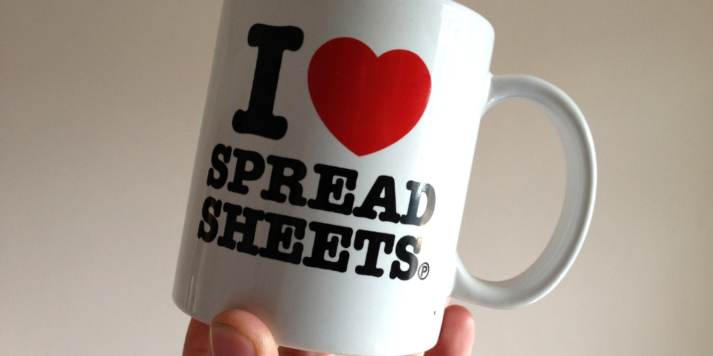 Someone holding a white the base of a mug that has 'I heart spreadsheets' printed on it.